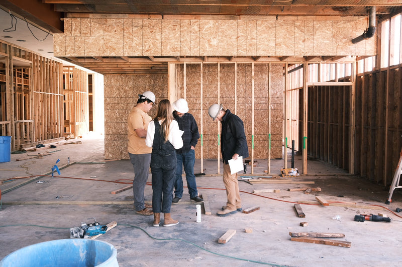 4 people, 3 in hard hats, review plans inside the wood-framed building during construction