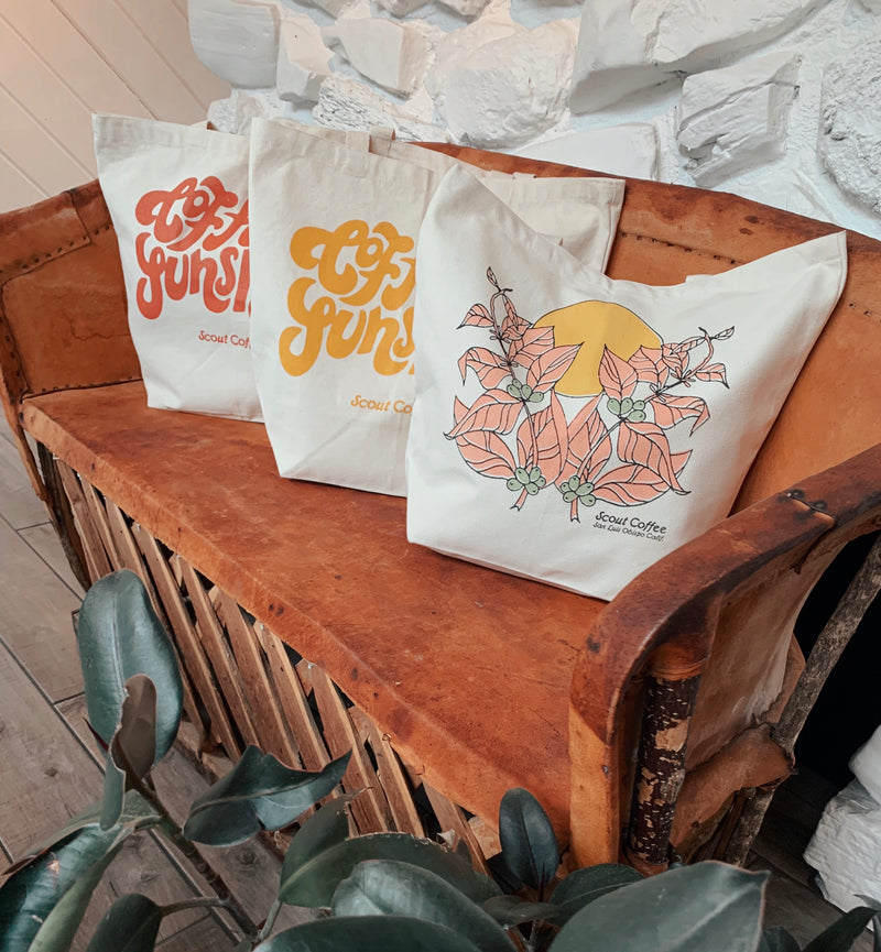 3 Scout Coffee tote bags, 2 Coffee and Sunshine prints and 1 floral print, on a worn leather bench