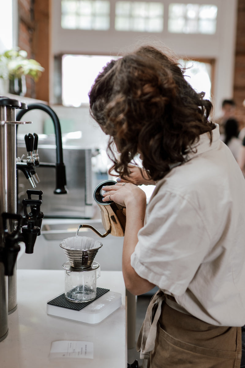 A barista pours water from a gooseneck kettle into a pour-over dripper on a scale