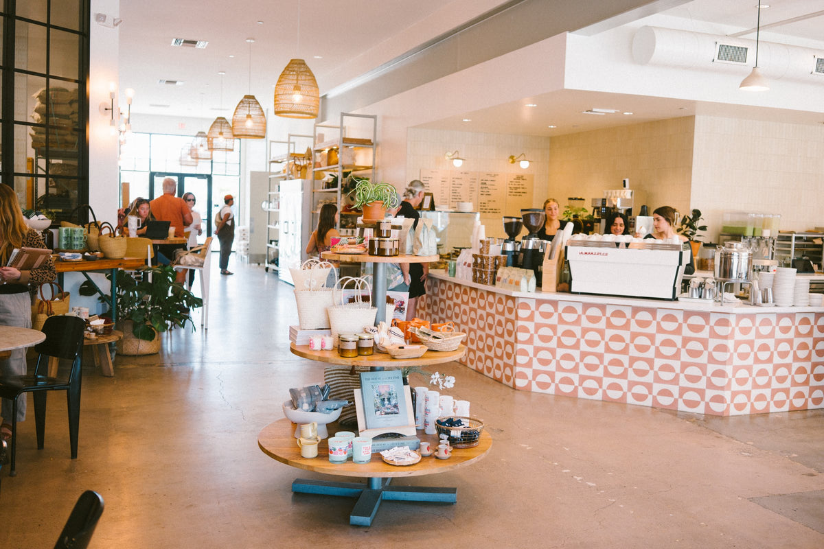 The Morro Bay Scout cafe with a round table of mugs and goods in front of the tiled coffee bar, baristas smiling behind the counter